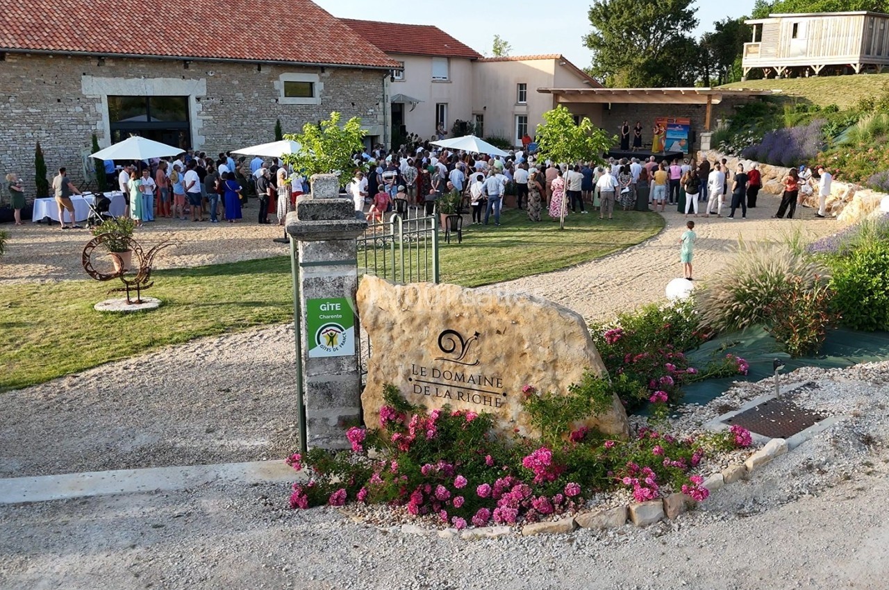 Vue d'un domaine avec des invités rassemblés en extérieur près d'un bâtiment en pierre et d'une enseigne fleurie.