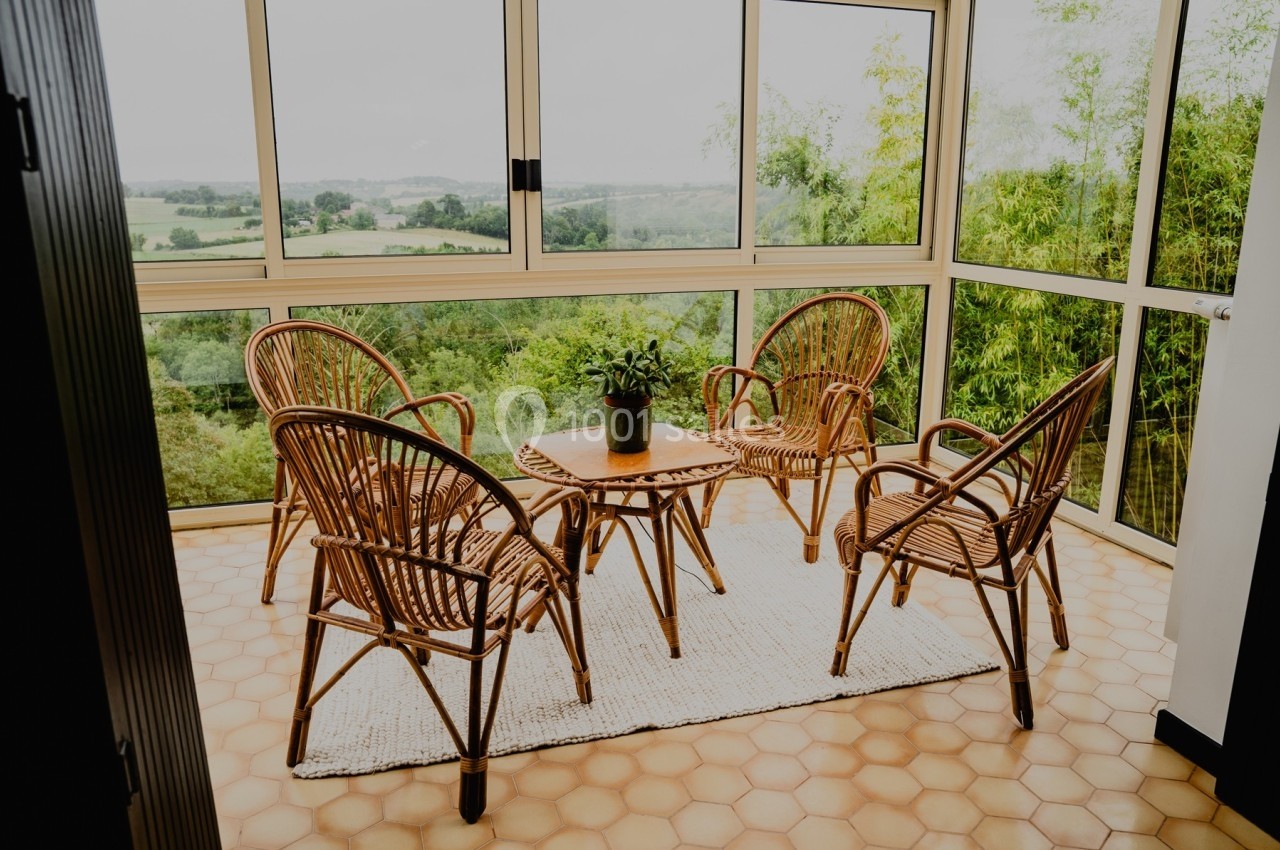 Quatre fauteuils en rotin autour d'une table basse sur un tapis clair dans une véranda avec vue sur la campagne.