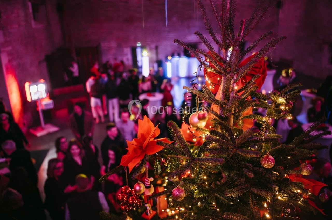 Sapin de Noël décoré avec lumières et ornements, surplombant une fête dans une salle éclairée chaleureusement.