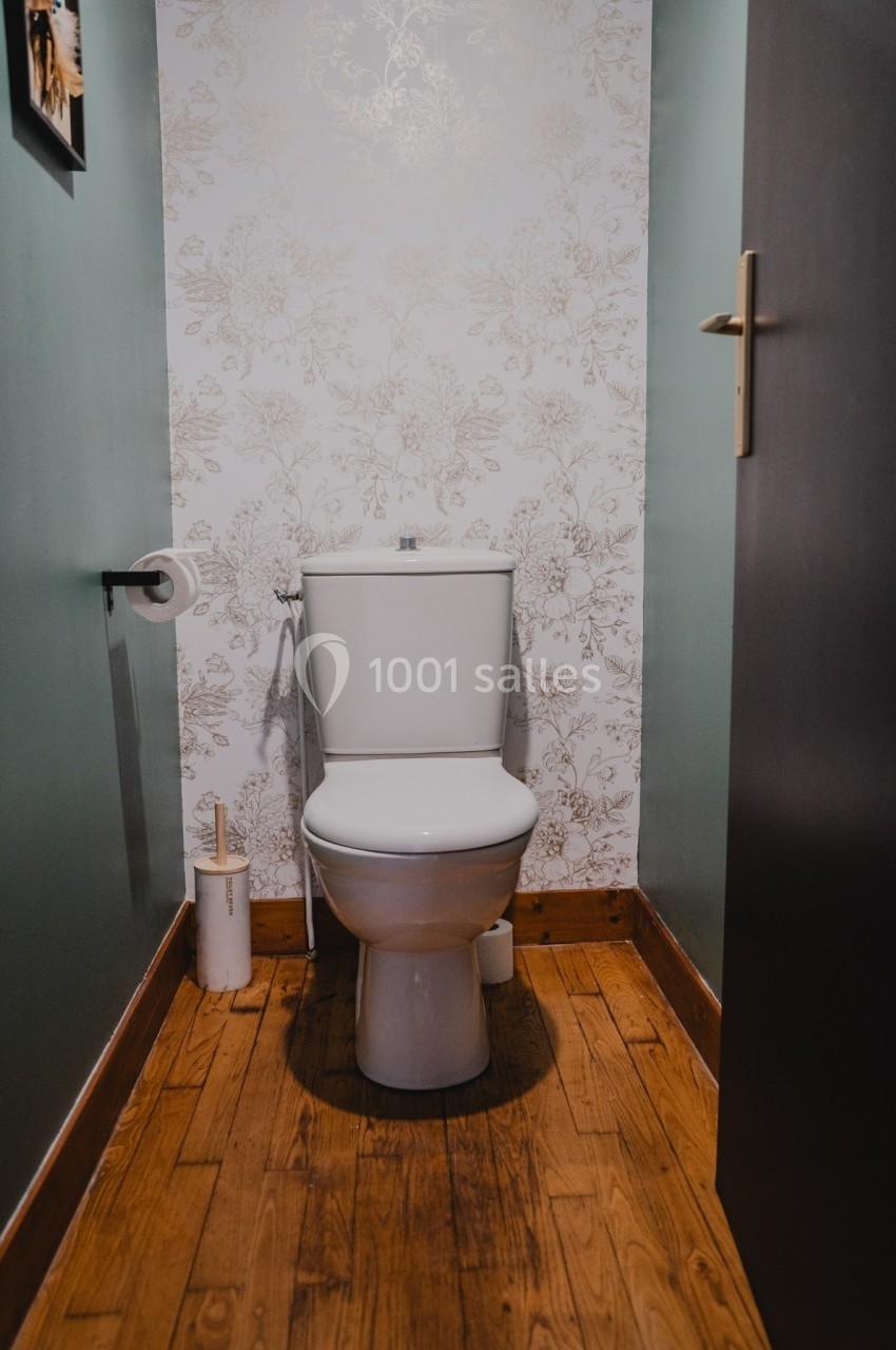 Toilettes avec réservoir blanc, papier toilette à gauche, brosse à droite, sol en bois et mur à motif floral.