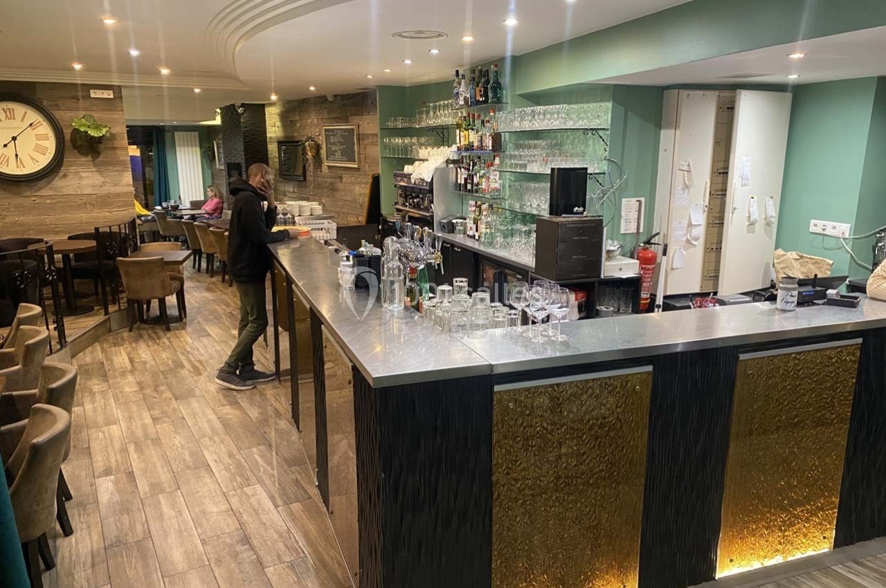 Un homme se tient derrière un comptoir de bar moderne dans un espace intérieur avec des tables et des chaises.