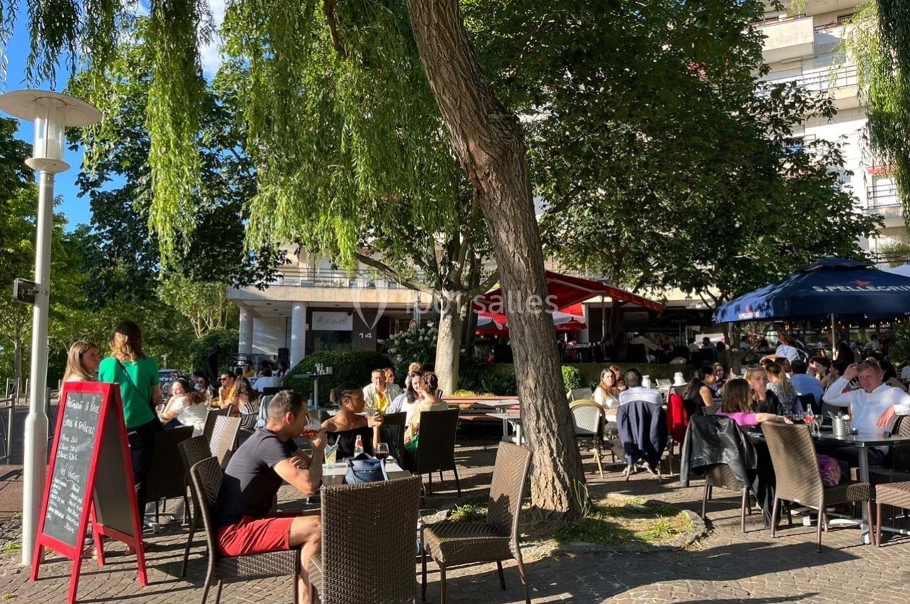 Terrasse d'un café-restaurant animée par des clients assis sous des arbres par une journée ensoleillée.