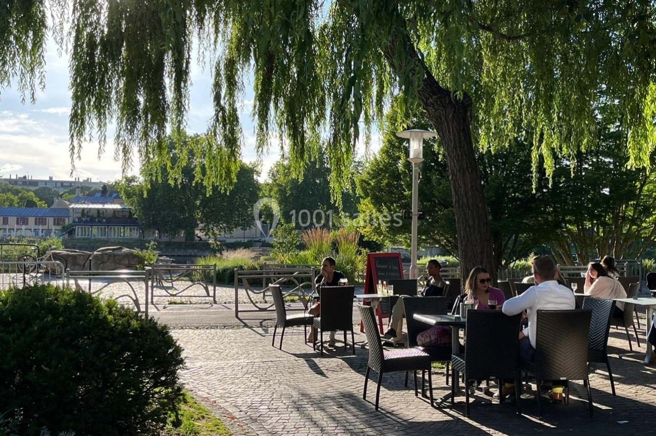 Des personnes assises à des tables en terrasse ombragée près d'un cours d'eau, entourées de verdure et de lumière naturelle.