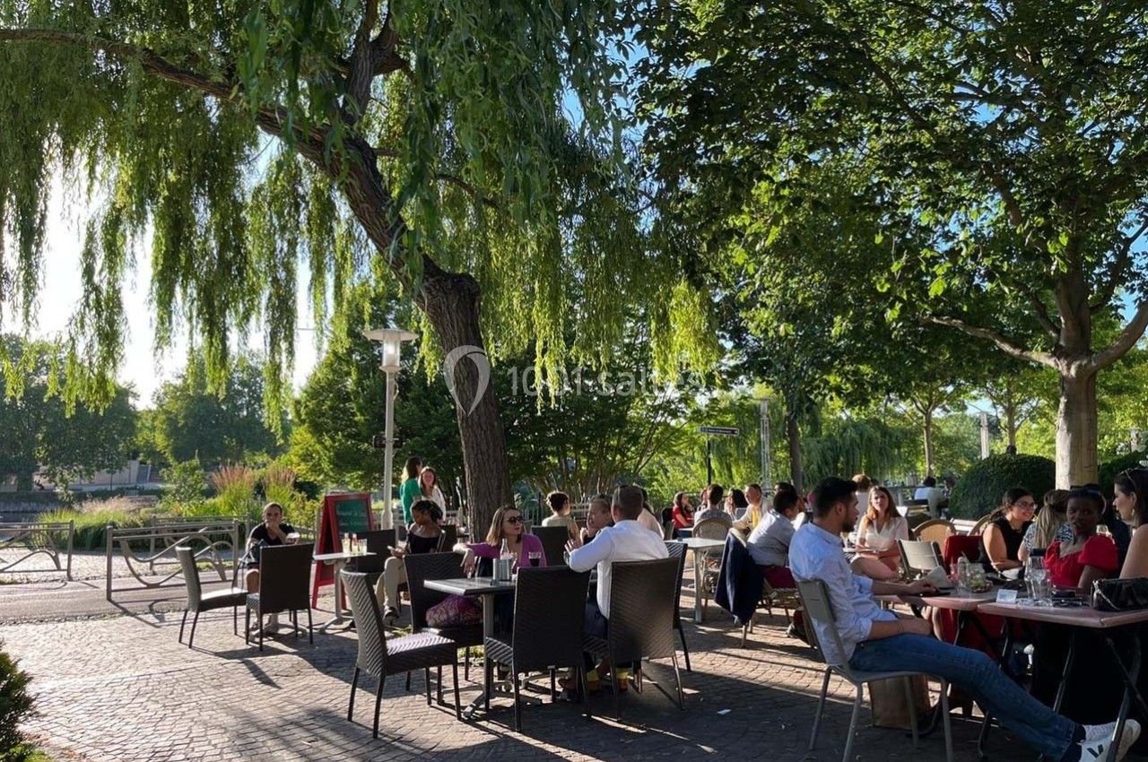 Des personnes assises en terrasse sous des arbres, profitant d'une journée ensoleillée près d'un espace vert.