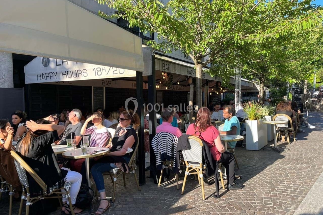 Terrasse de café animée par une journée ensoleillée, avec des clients assis autour de tables sur une rue pavée.