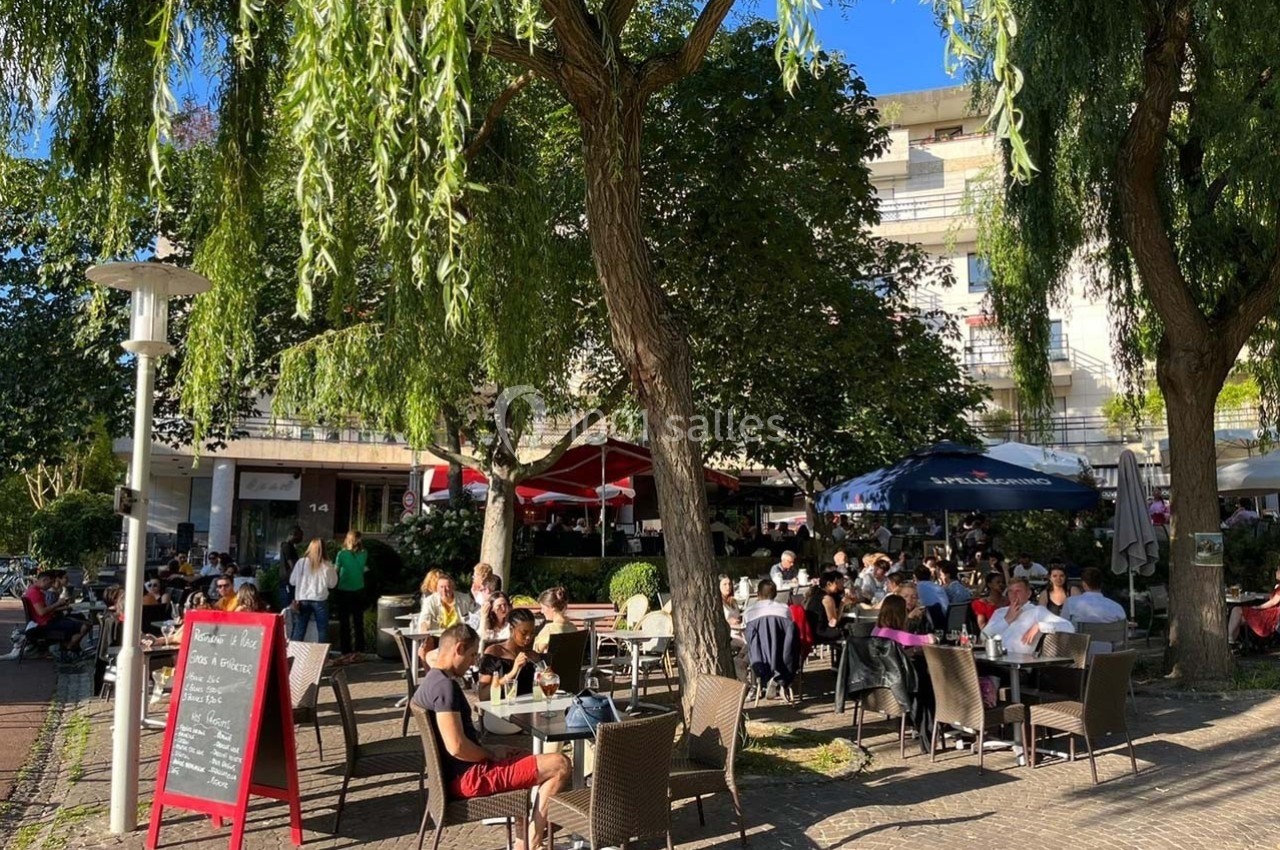 Terrasse d'un café animée avec des clients assis sous des arbres, par une journée ensoleillée.