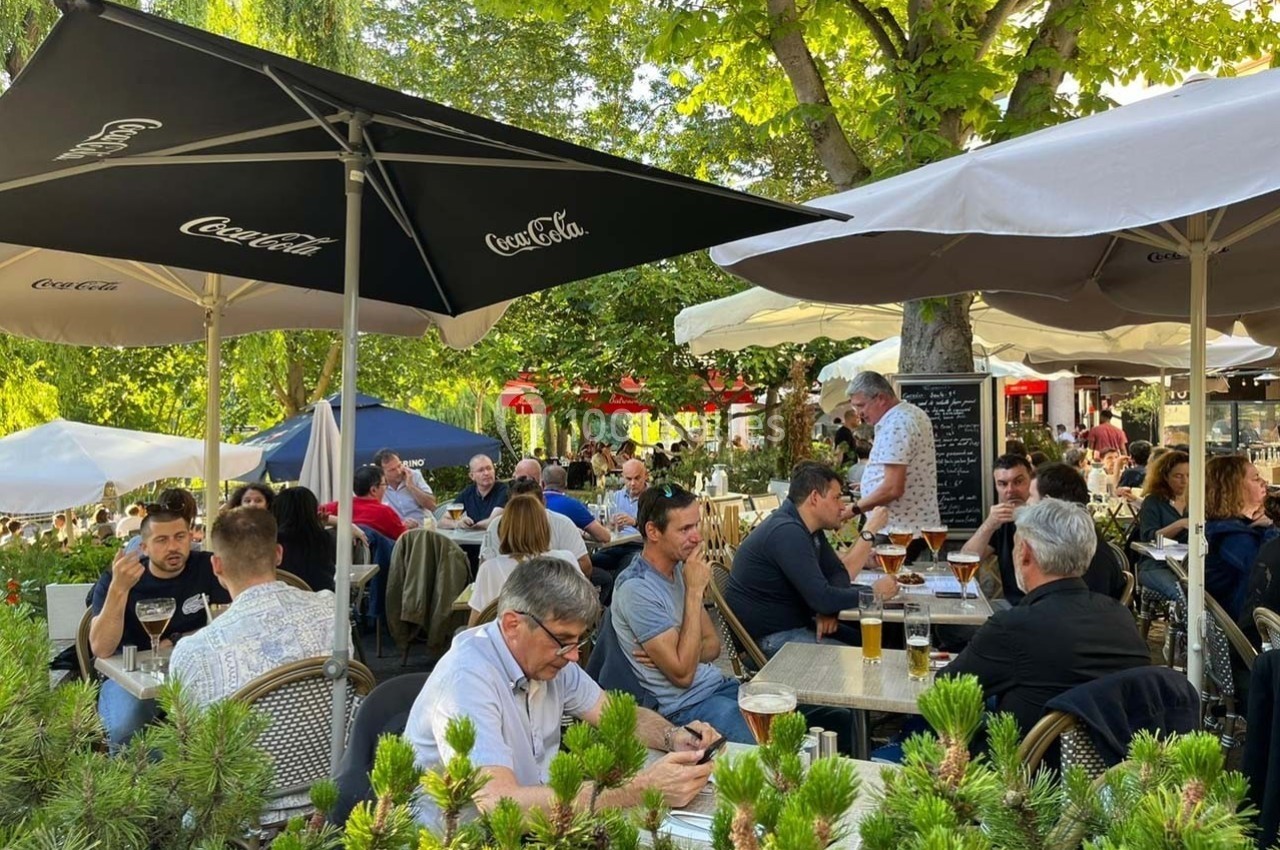 Terrasse animée d'un café en plein air, avec des clients attablés sous des parasols, entourés de verdure.