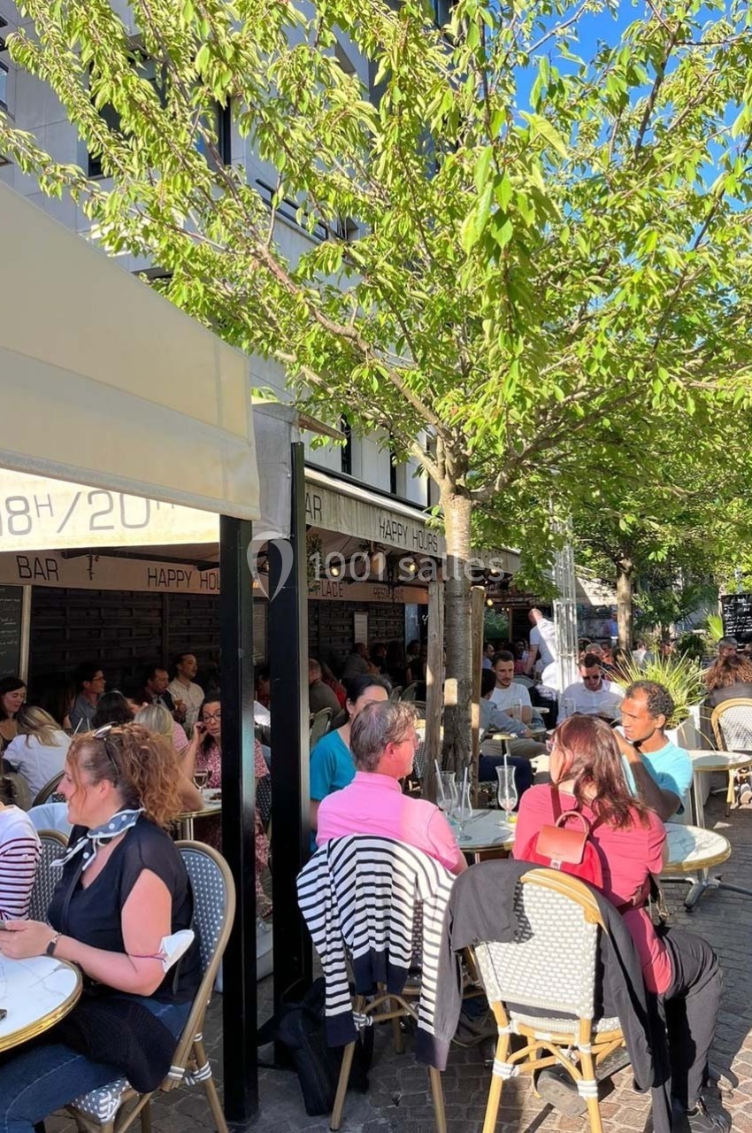 Terrasse animée d'un café sous un arbre, avec des clients assis à des tables en plein air par une journée ensoleillée.