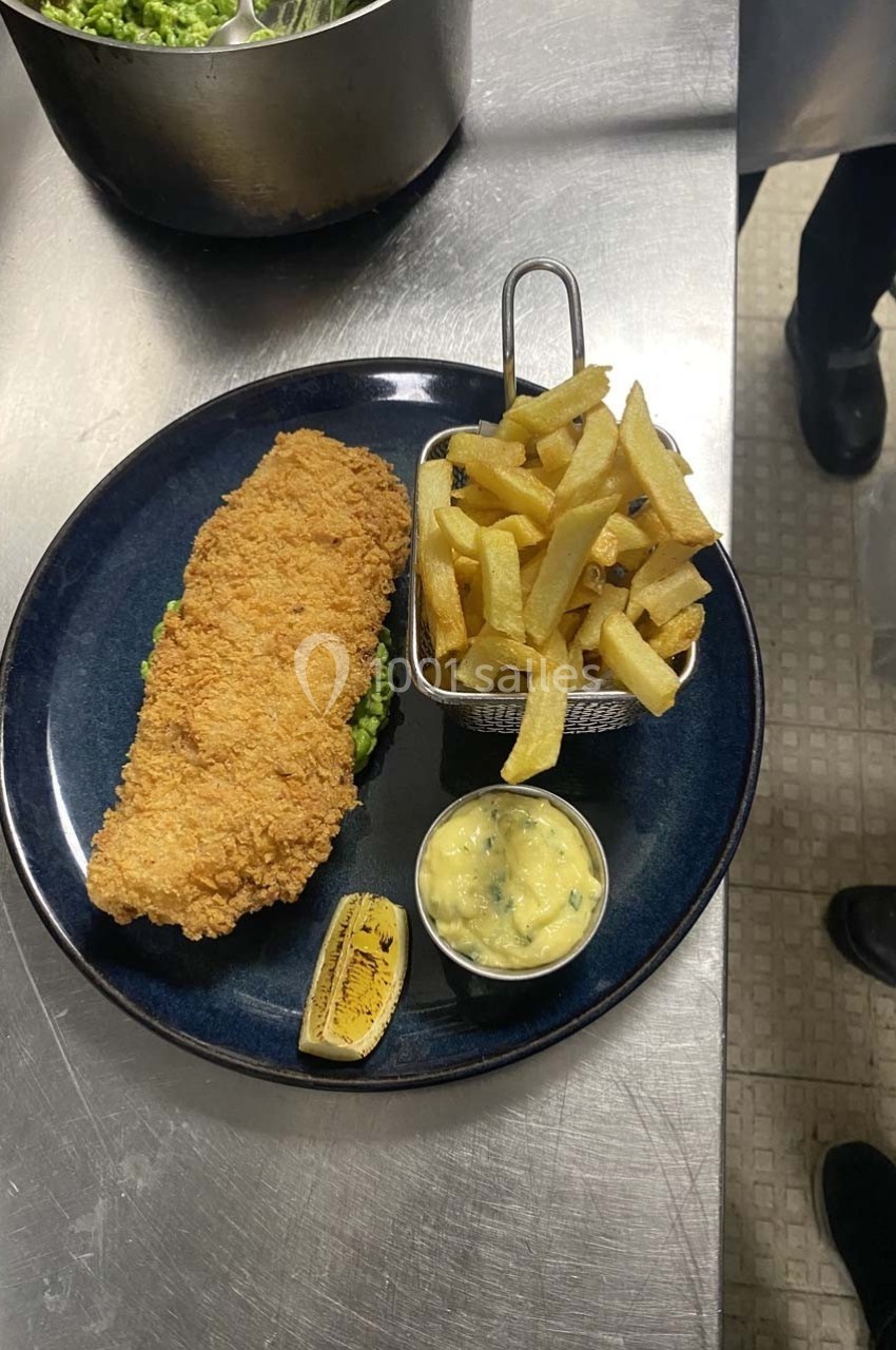 Assiette avec un filet de poisson pané, des frites dans un panier, purée de pois, rondelle de citron grillé et sauce.
