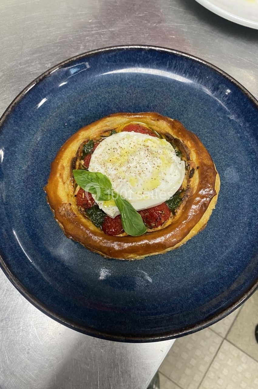 Tartelette garnie de tomates confites, pesto, œuf poché et feuille de basilic, servie sur une assiette bleue.