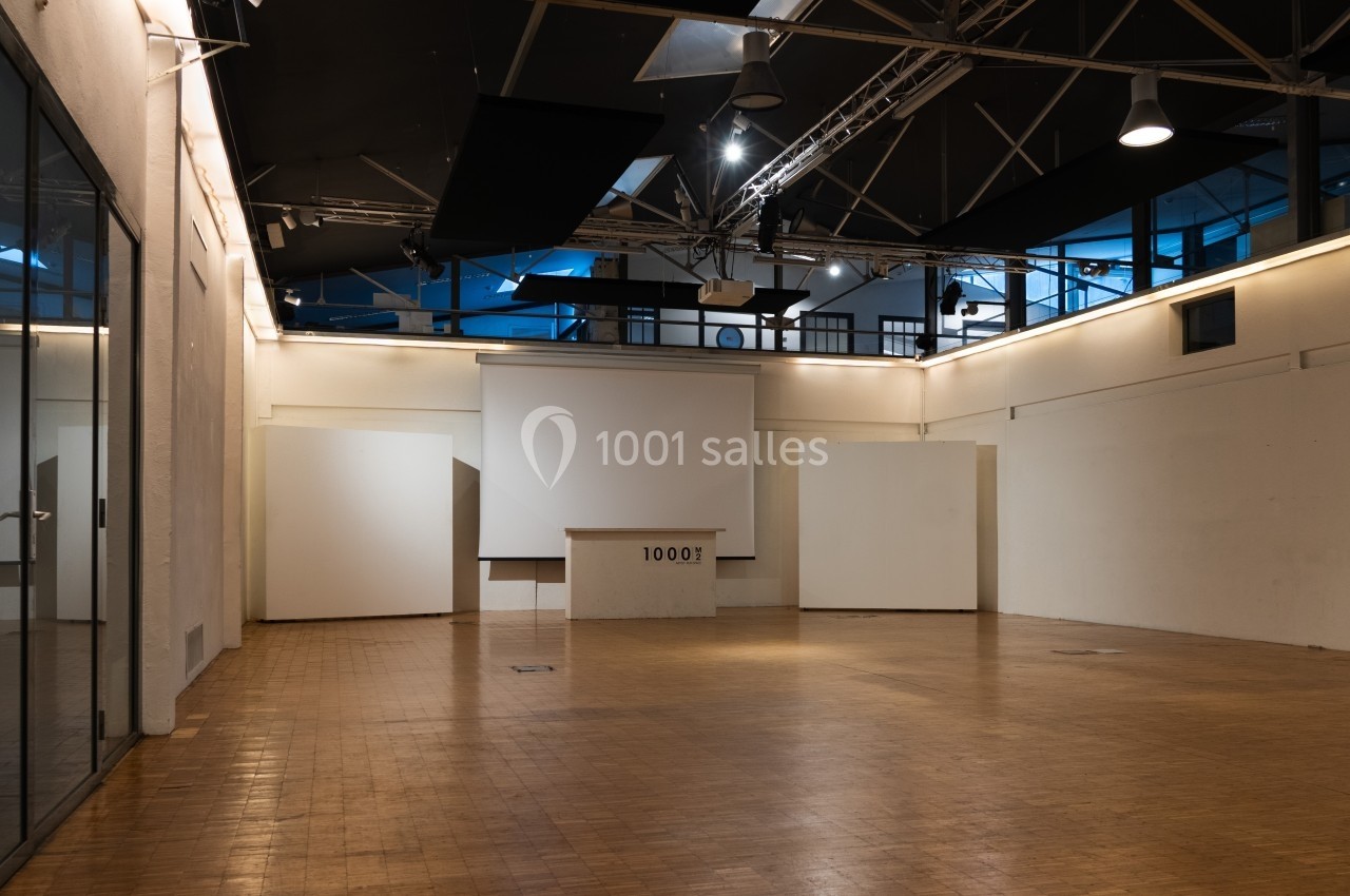 Salle vide avec parquet en bois, murs blancs, plafond industriel et écran de projection au fond.