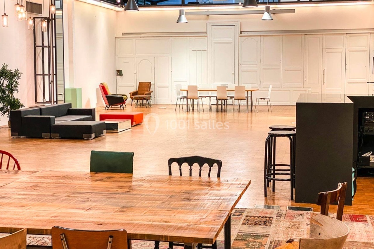 Salle spacieuse avec parquet, mobilier varié, tables en bois, canapés et chaises, éclairée par des lumières suspendues.