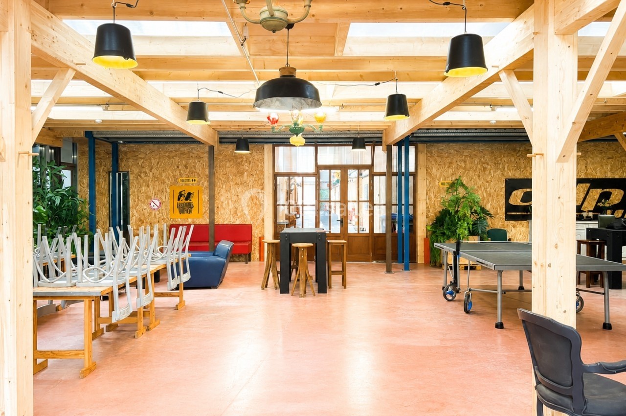 Salle lumineuse avec structure en bois, tables, chaises empilées, table de ping-pong et plantes décoratives.