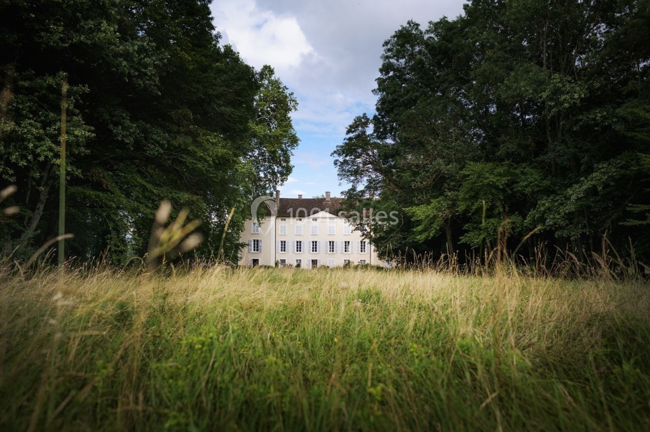 Manoir blanc entouré d'arbres, vu à travers une prairie verdoyante sous un ciel partiellement nuageux.