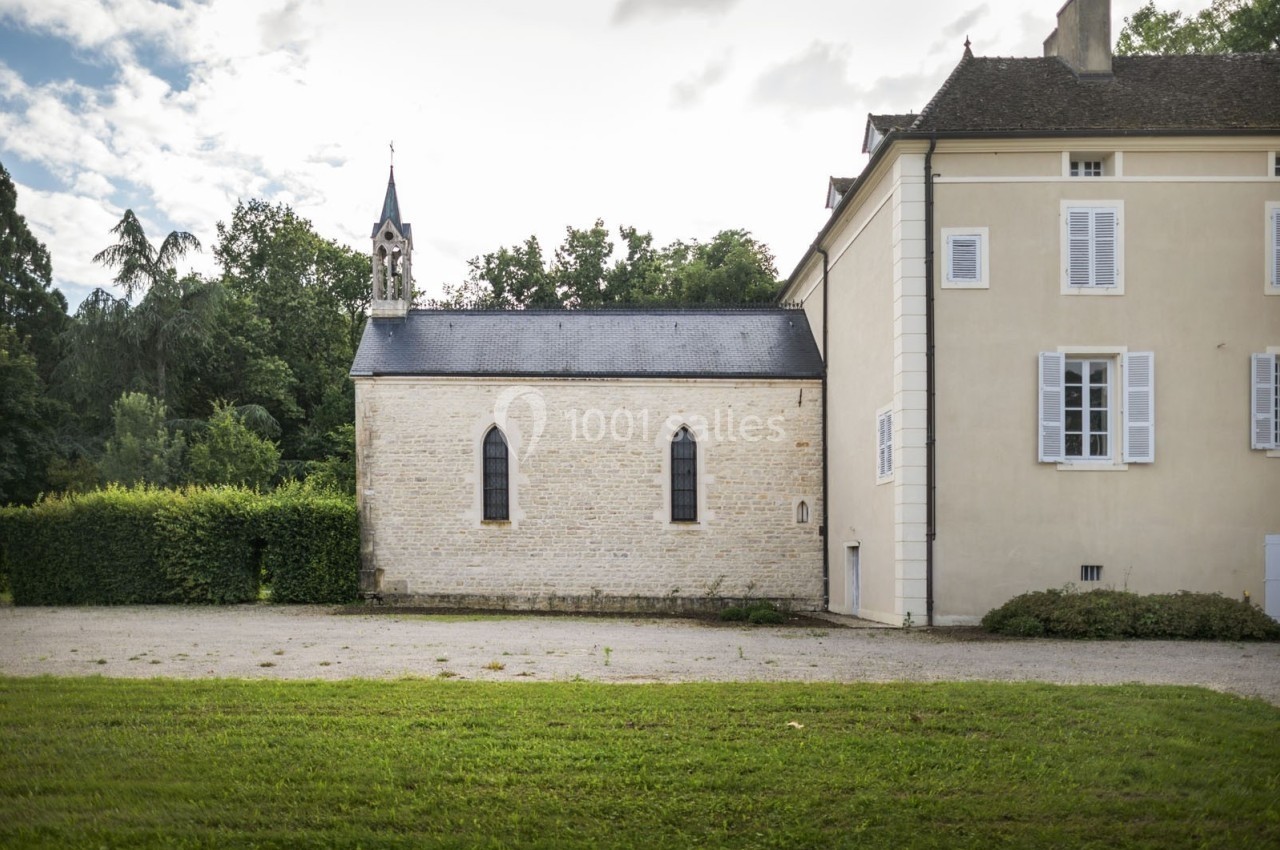 Petite chapelle en pierre attenante à un bâtiment ancien, entourée de verdure sous un ciel partiellement nuageux.