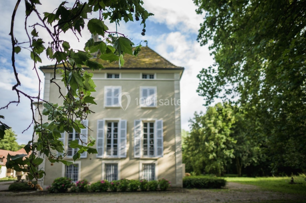 Façade d'une maison ancienne beige avec volets blancs, entourée de verdure sous un ciel partiellement nuageux.