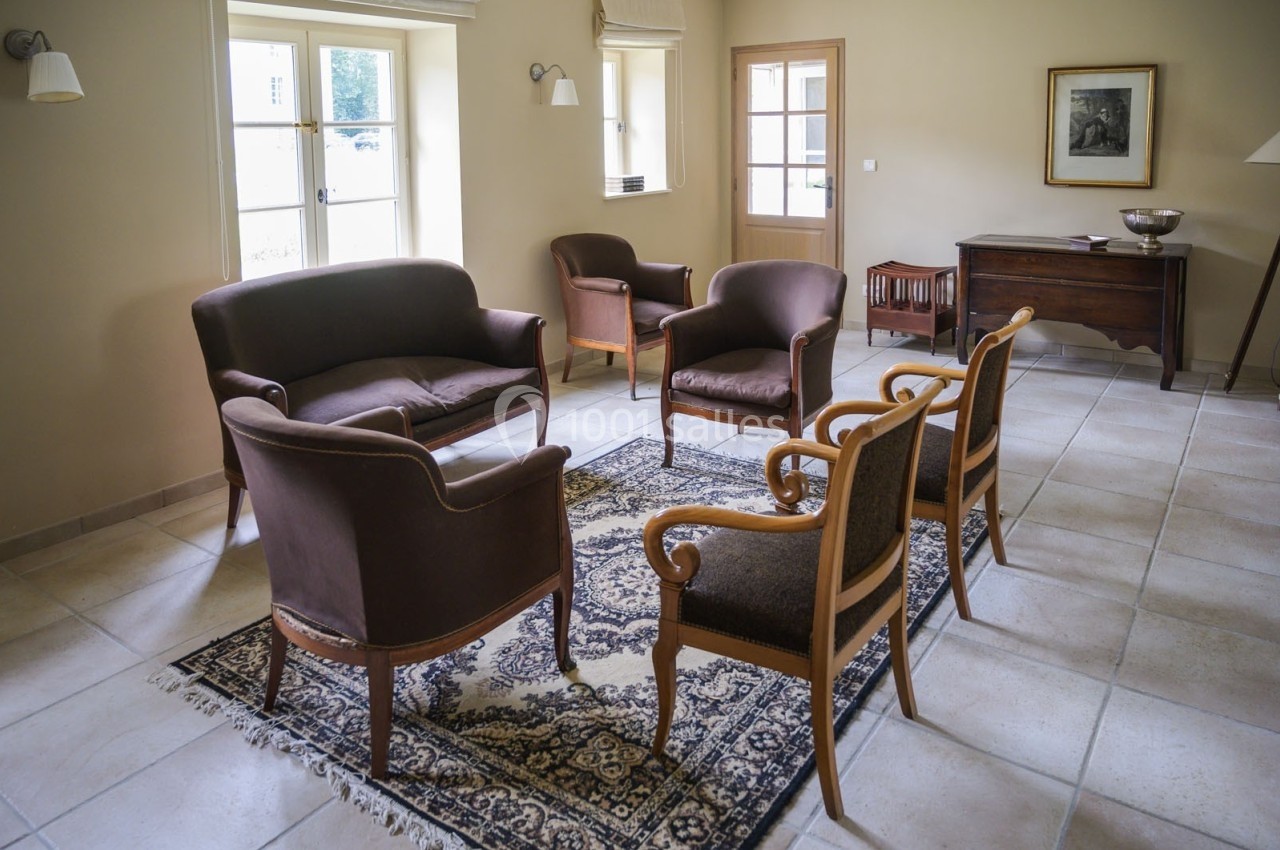 Salon avec fauteuils et canapé en bois et tissu, disposés autour d'un tapis, éclairé par des fenêtres à double battant.