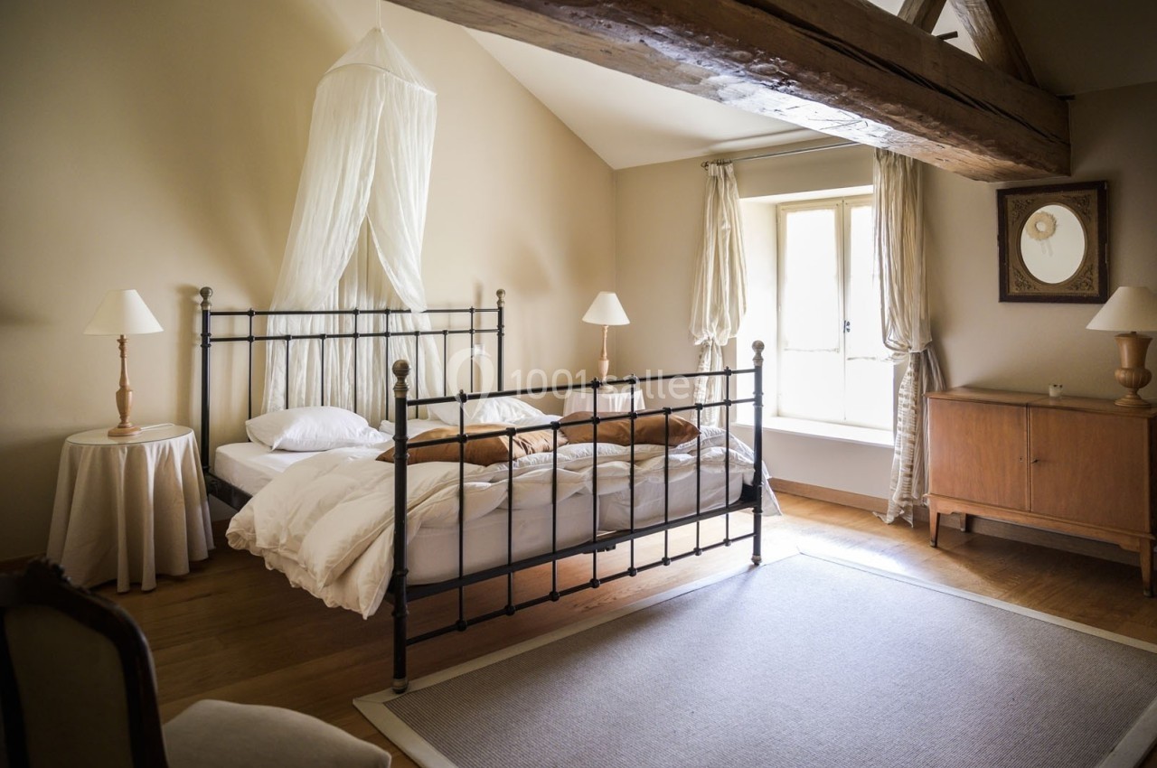 Chambre lumineuse avec lit à baldaquin en métal, poutre apparente, mobilier en bois et grande fenêtre.