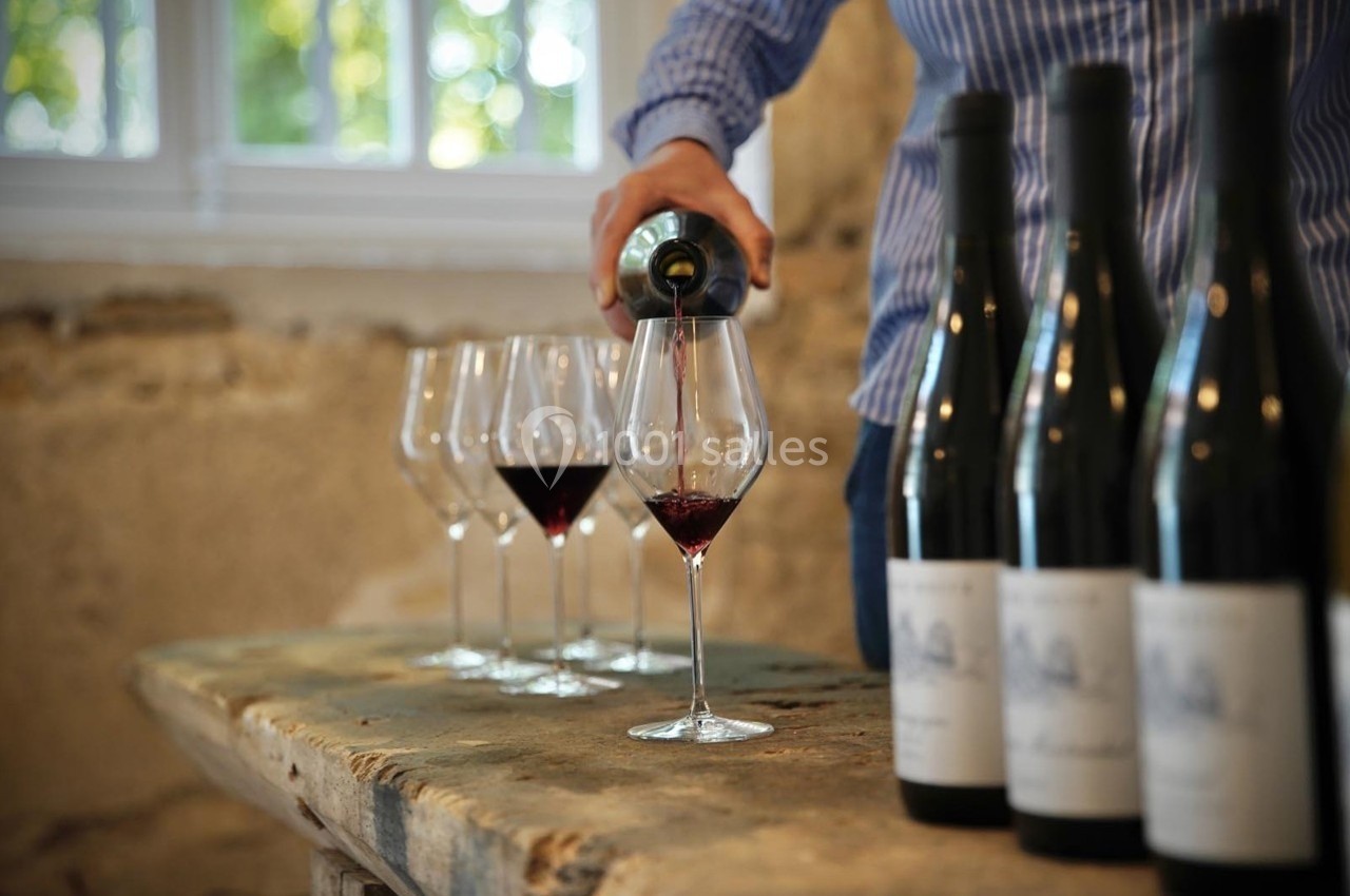 Une personne verse du vin rouge dans un verre sur une table en bois, avec des bouteilles et des verres alignés.