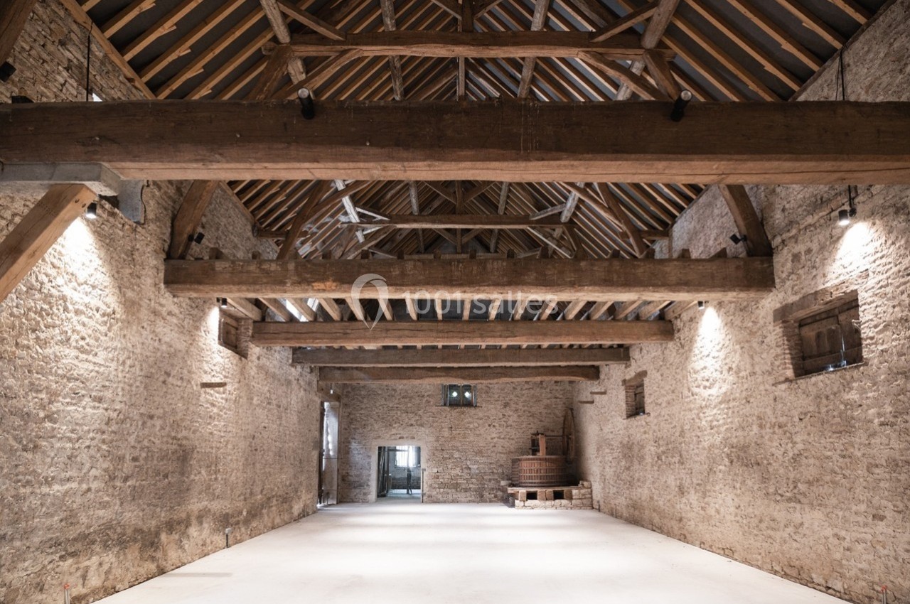 Intérieur d'une grande grange en pierre avec poutres en bois apparentes et sol clair.
