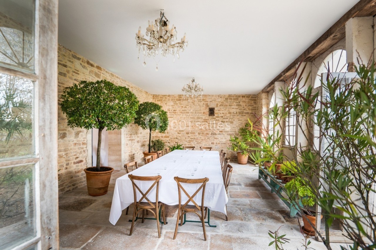 Salle lumineuse avec table rectangulaire dressée, chaises en bois, murs en pierre, plantes en pot et lustres suspendus.