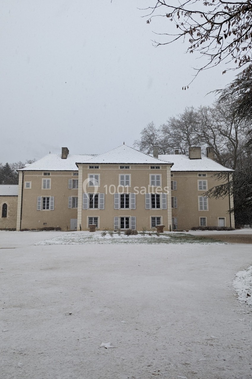 Façade d'un bâtiment ancien beige avec toit enneigé, entouré d'arbres en hiver.