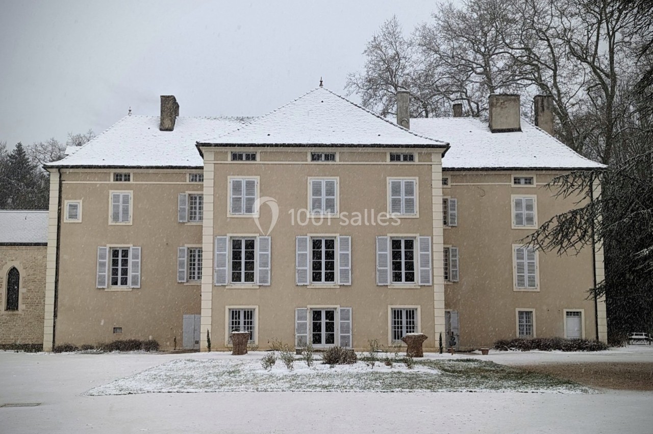 Façade d'un grand bâtiment ancien beige avec des volets blancs, entouré de neige et d'arbres en hiver.