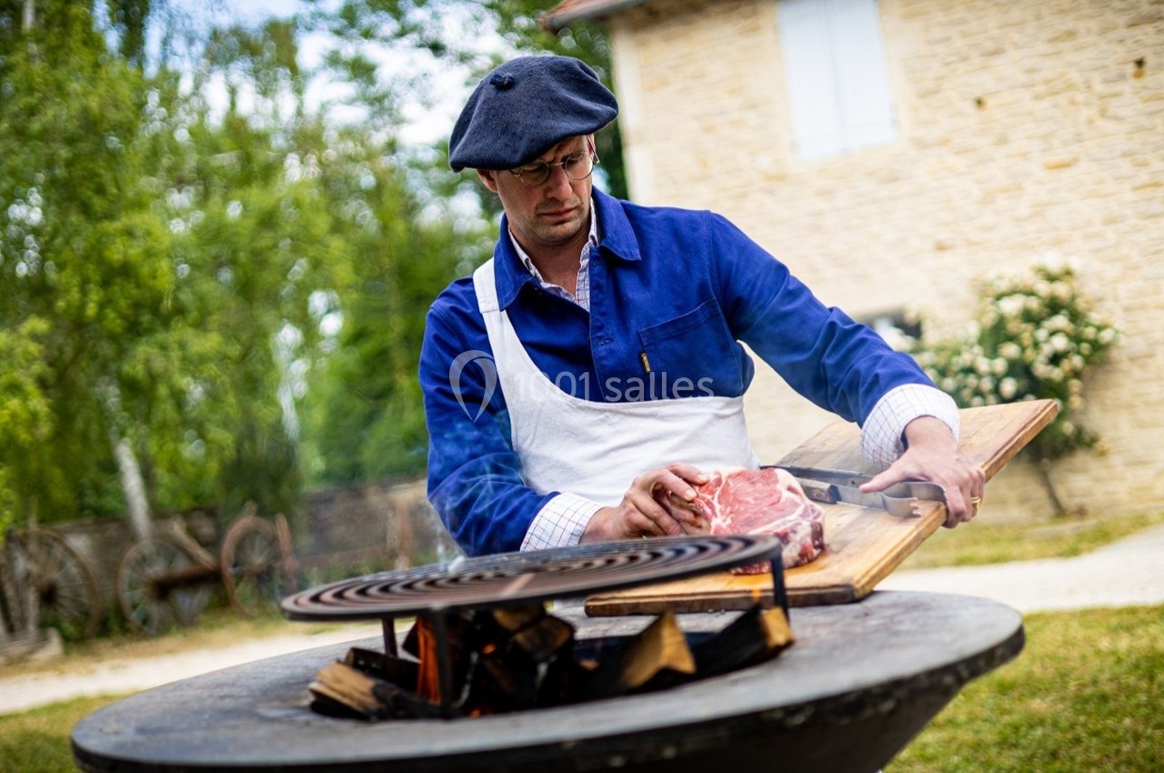 Homme en tablier préparant un morceau de viande sur une planche près d'un barbecue en plein air, devant une maison en pierre.