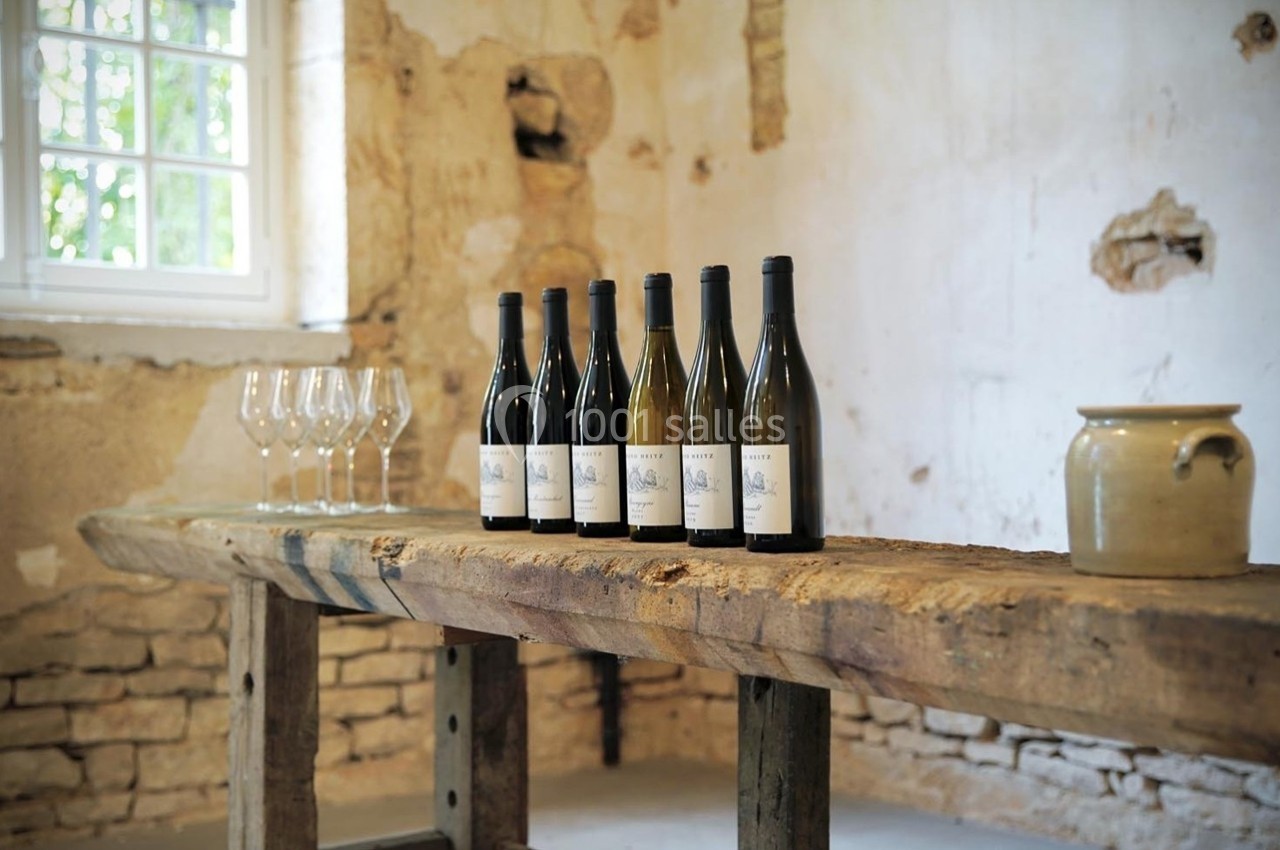 Bouteilles de vin alignées sur une table en bois rustique, avec des verres et un pot en céramique dans une pièce en pierre.