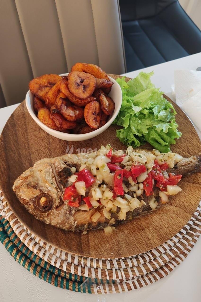 Poisson grillé garni de légumes, accompagné de bananes plantains frites et de salade verte sur une assiette en bois.