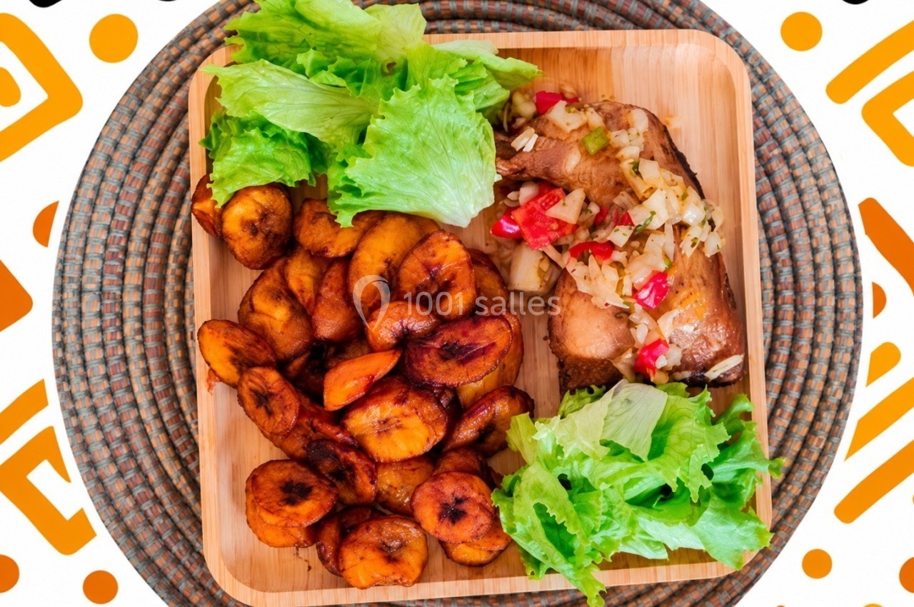 Assiette en bois contenant des rondelles de plantains frits, du poisson garni de légumes et des feuilles de salade verte.