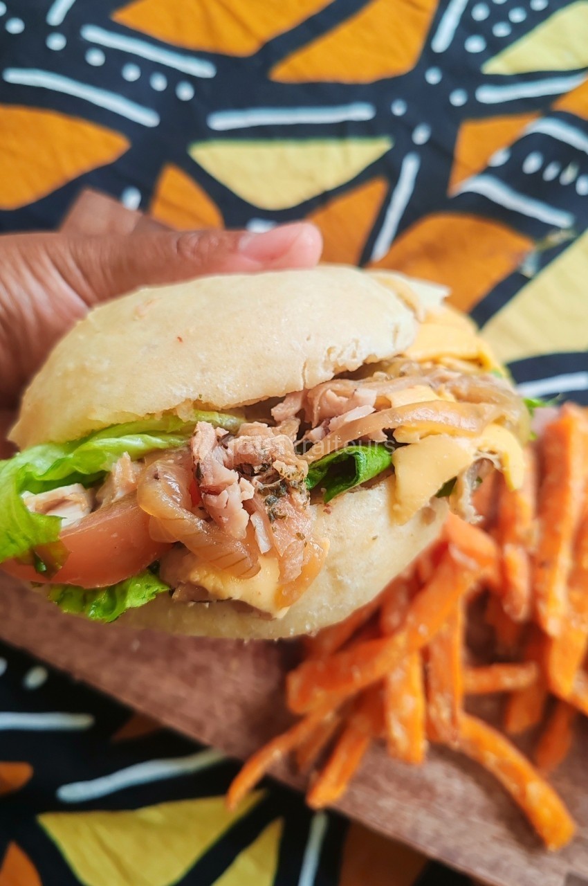 Sandwich garni de légumes, fromage et viande, tenu en main, accompagné de frites de patate douce sur une table colorée.