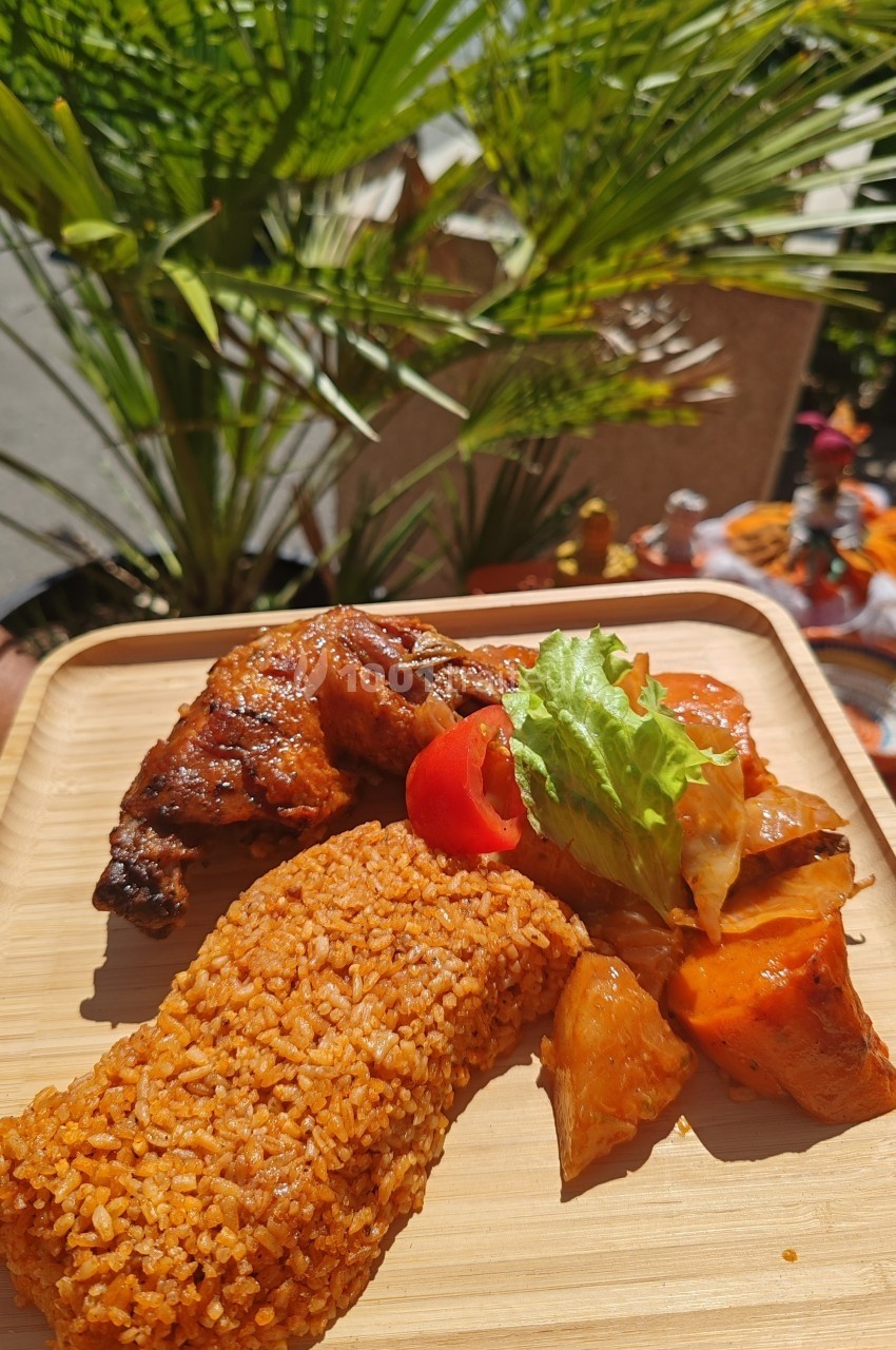 Assiette en bois contenant du riz rouge, du poulet grillé, des légumes et une garniture de salade, en extérieur ensoleillé.