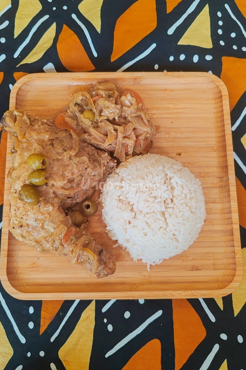 Assiette en bois contenant du riz blanc et des morceaux de poulet en sauce avec des olives, sur une nappe colorée.