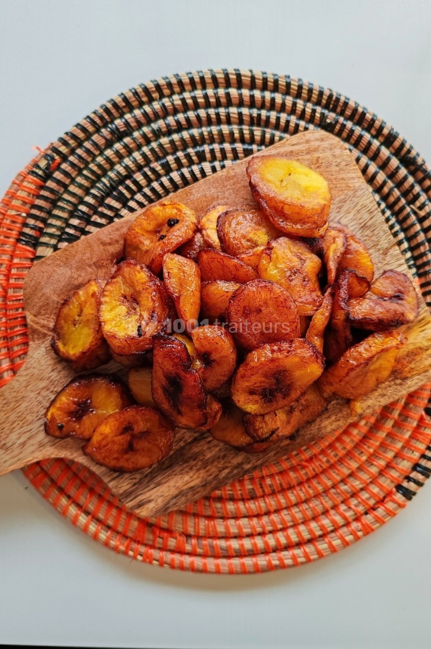 Tranches de bananes plantains frites disposées sur une planche en bois, posée sur un dessous de plat coloré.