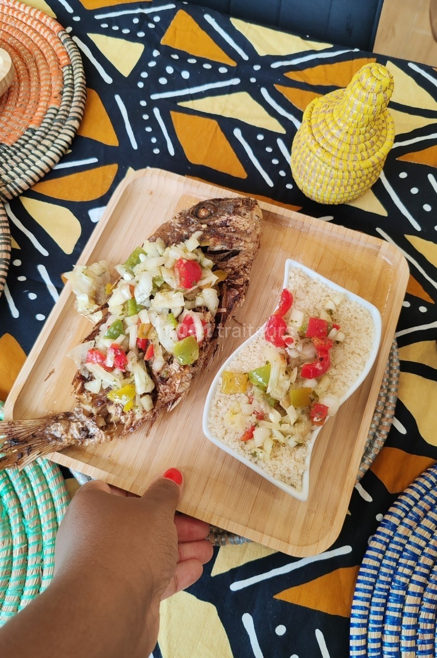 Poisson grillé garni de légumes colorés accompagné de semoule dans un plat sur une table au motif africain.