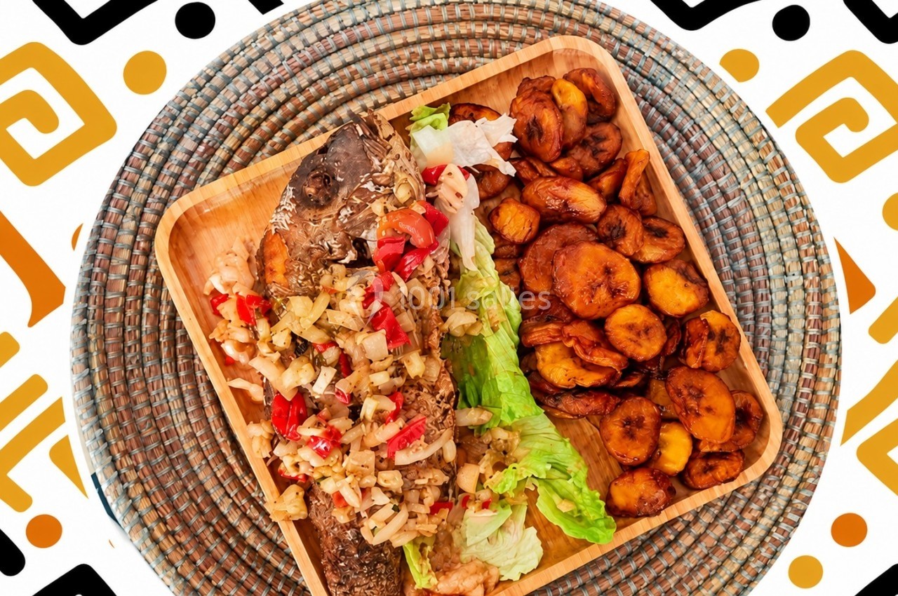 Plat de poisson grillé garni de légumes, accompagné de bananes plantains frites et de laitue, sur un plateau en bois.