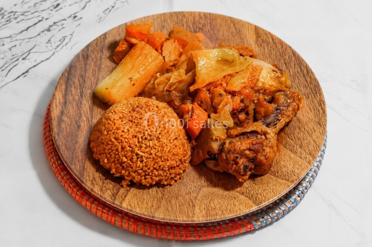 Assiette en bois contenant du poulet rôti, légumes mijotés et boule de couscous rouge, posée sur une table blanche.