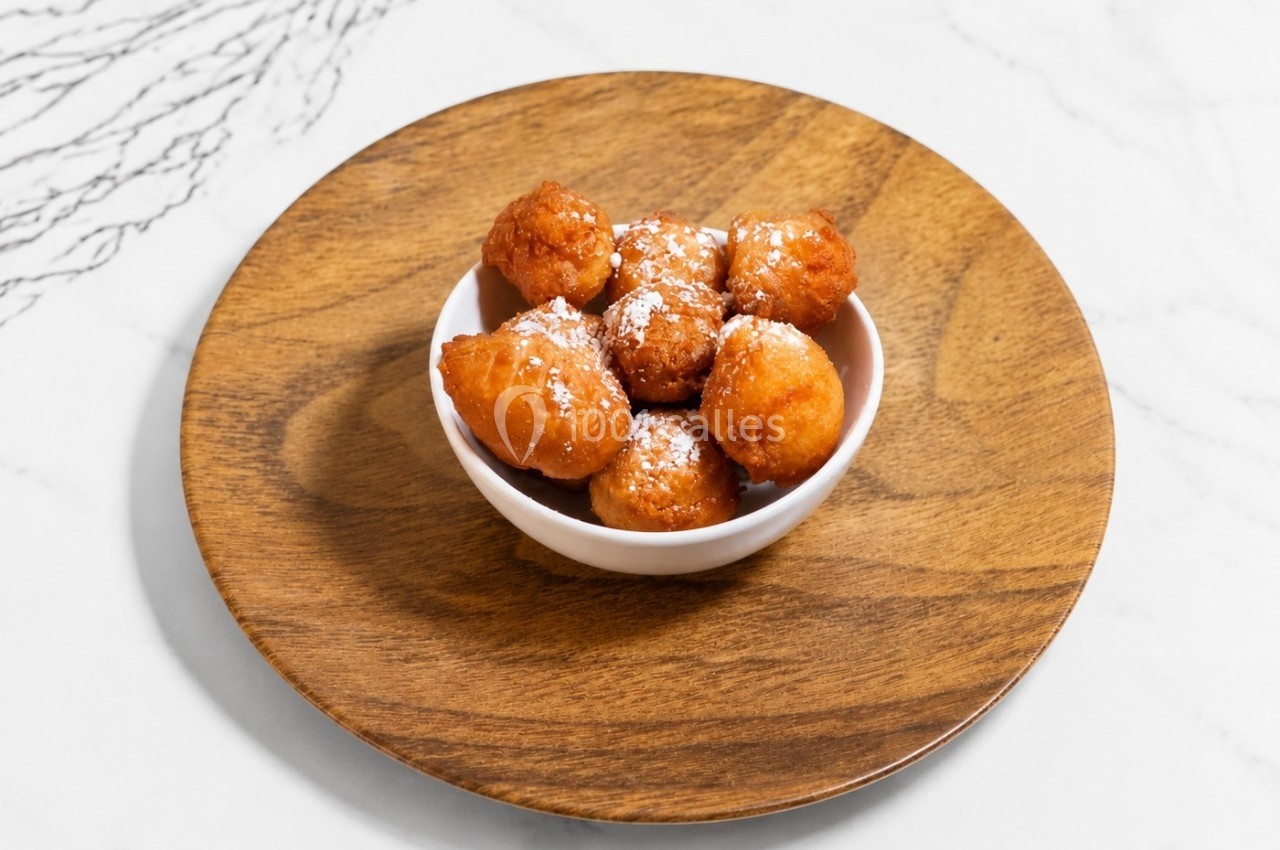 Beignets dorés saupoudrés de sucre glace, servis dans un petit bol blanc sur une assiette en bois.