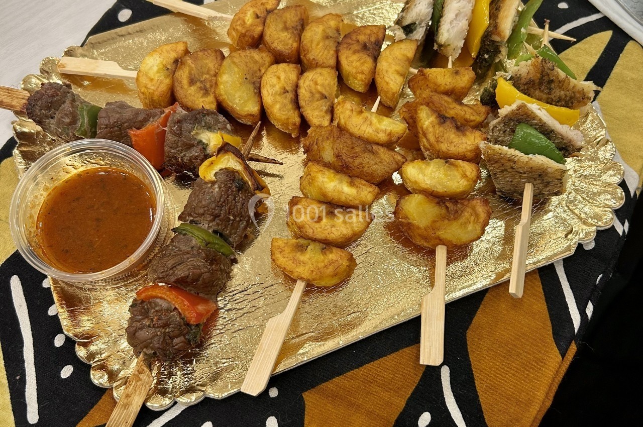 Assortiment de brochettes de viande, légumes, poisson et plantains frits, présenté sur un plateau doré avec une sauce.