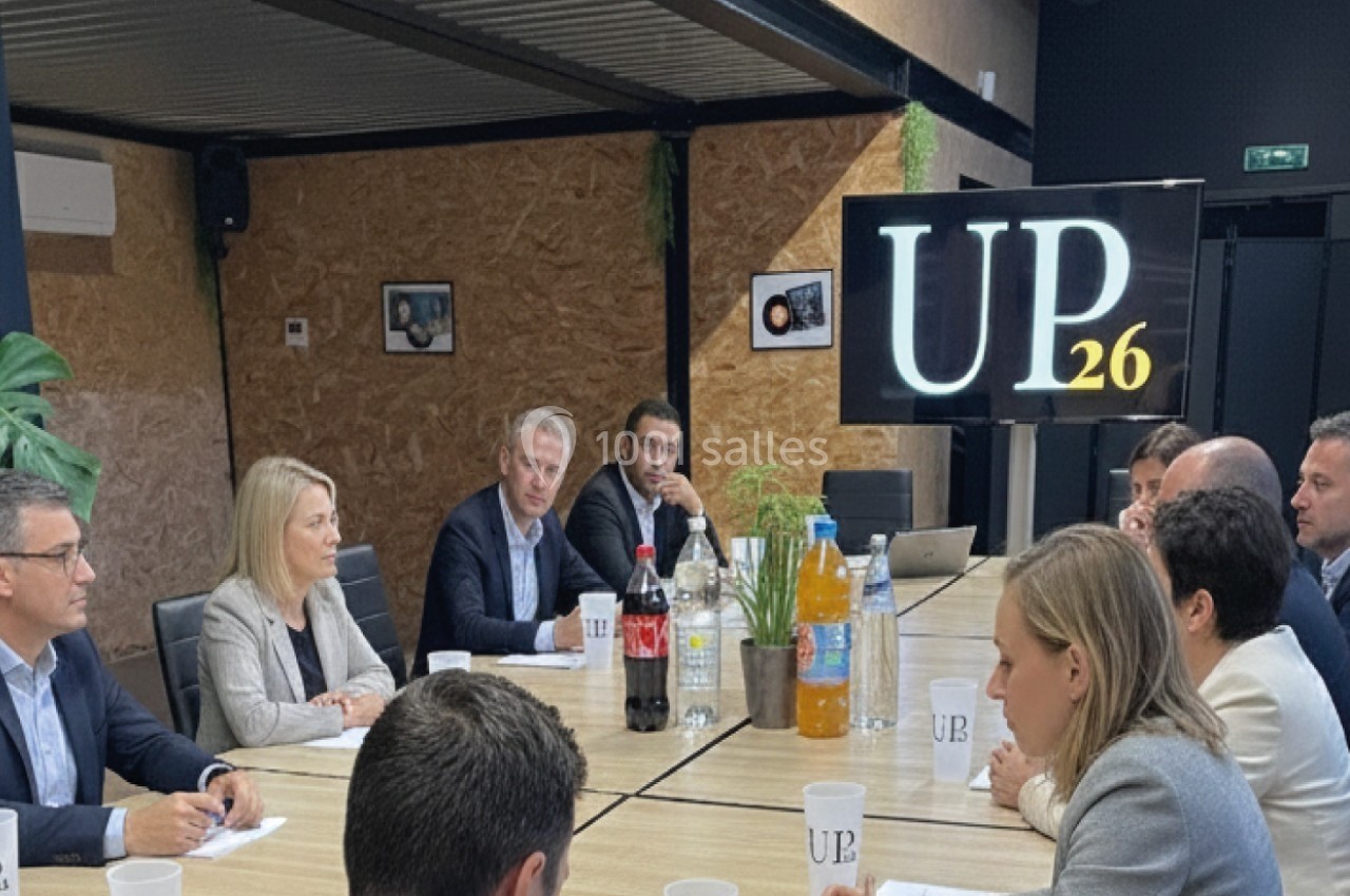 Des personnes discutent autour d'une grande table de réunion, avec des boissons et un écran affichant ’UP26’.