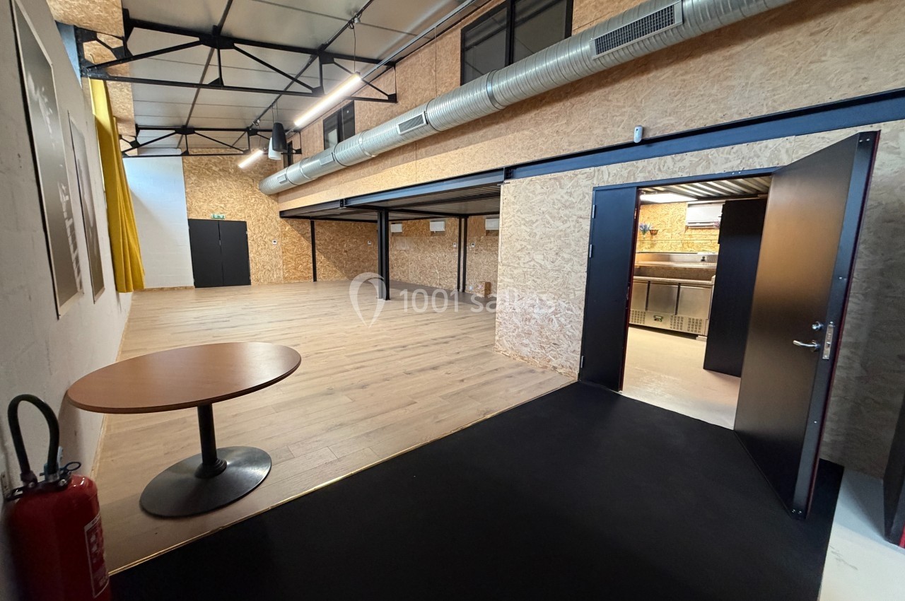 Espace intérieur avec sol en bois, murs en OSB, mezzanine, table ronde, extincteur et porte ouverte sur une cuisine.