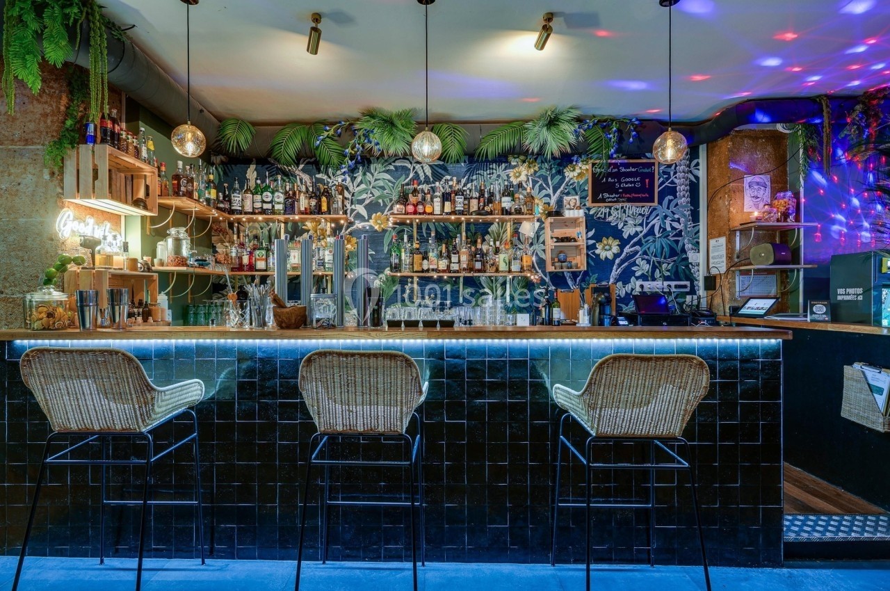 Bar avec comptoir en carrelage noir, tabourets en rotin, étagères remplies de bouteilles et décoration végétale.