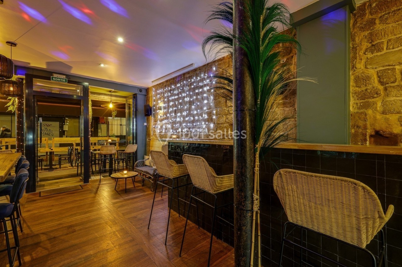 Intérieur d'un bar avec des chaises en rotin, un mur en pierre éclairé par des guirlandes lumineuses et un sol en bois.
