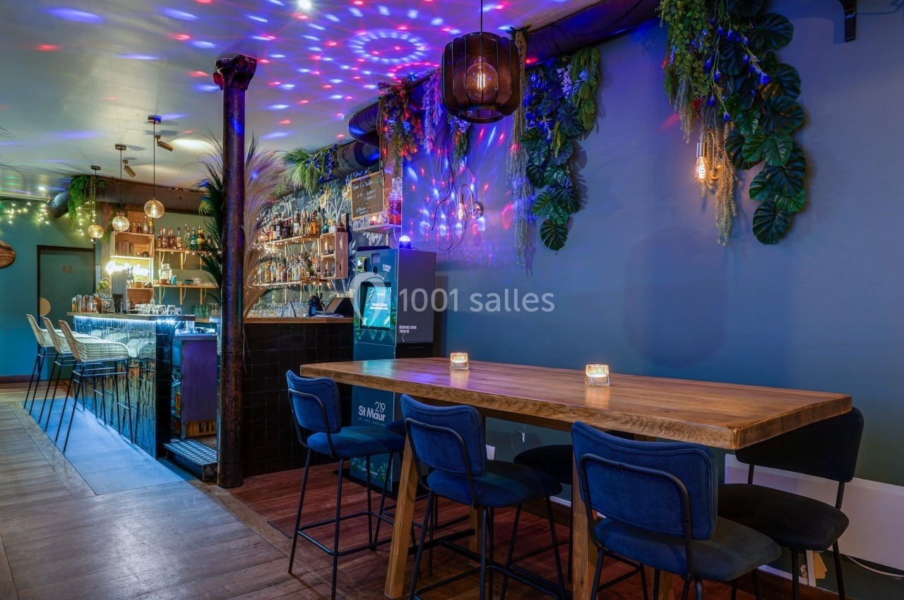 Salle de bar avec table en bois, chaises bleues, éclairage coloré et décorations végétales sur un mur bleu.