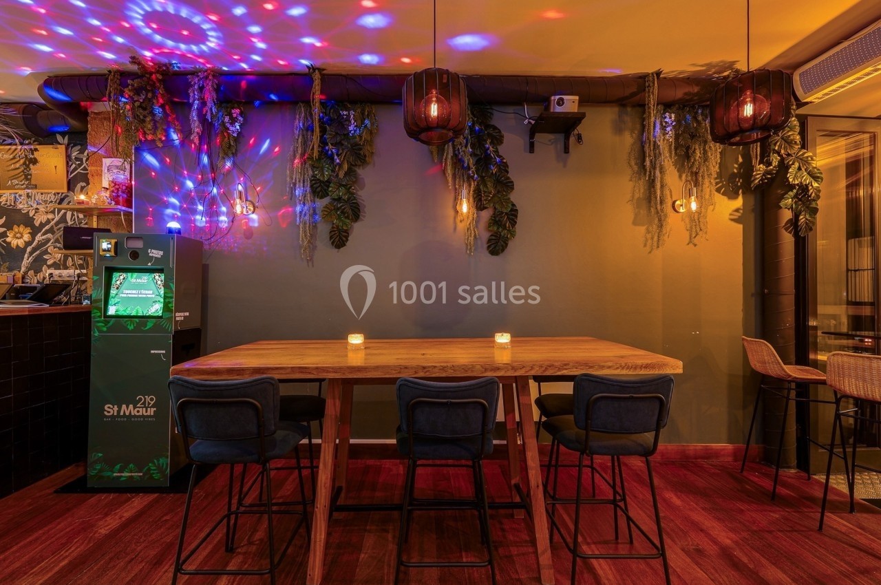 Salle avec table en bois, chaises hautes, éclairage tamisé, décorations végétales suspendues et lumières colorées.