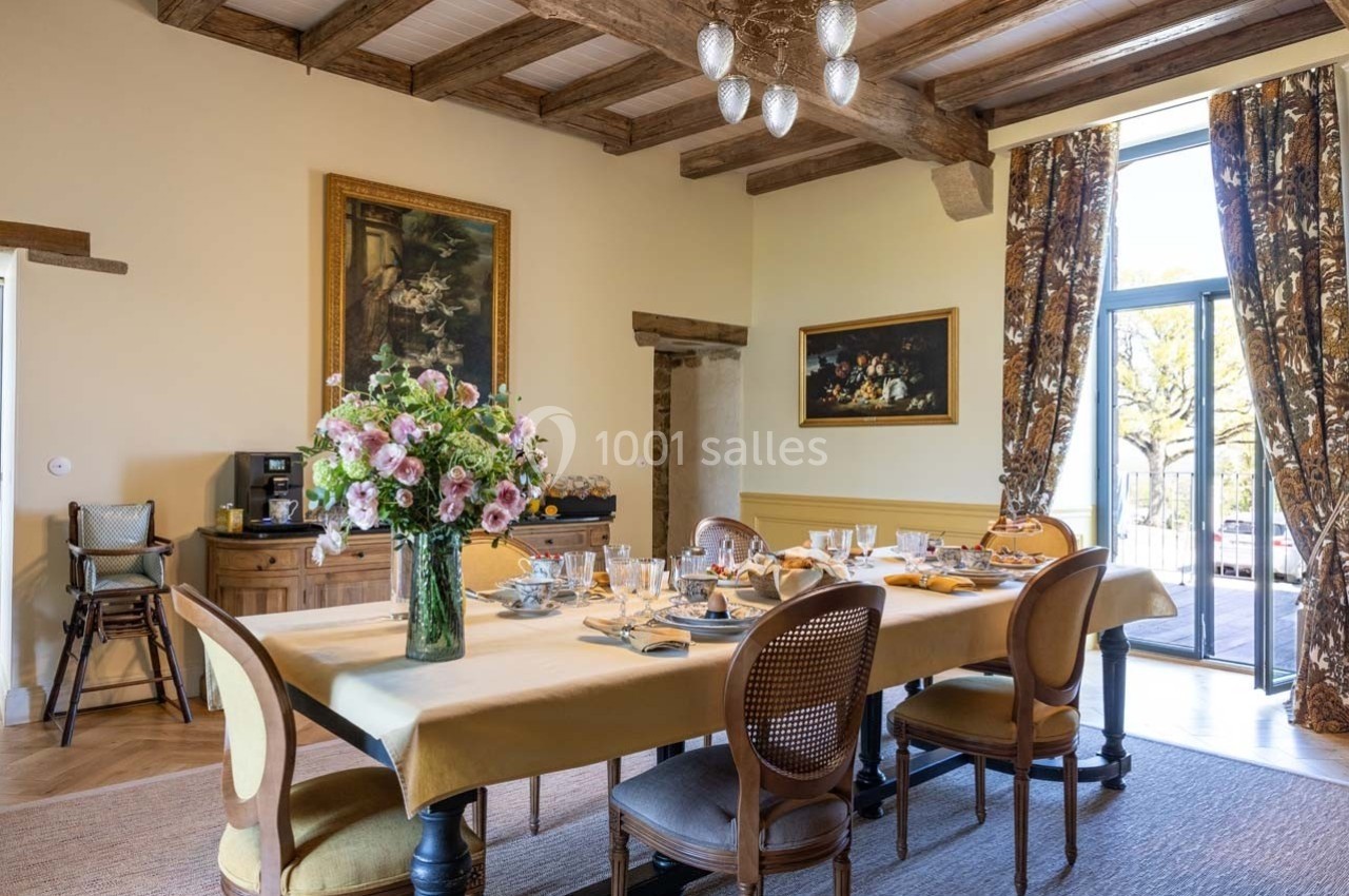 Salle à manger lumineuse avec une grande table dressée, bouquet de fleurs au centre et vue sur l'extérieur.