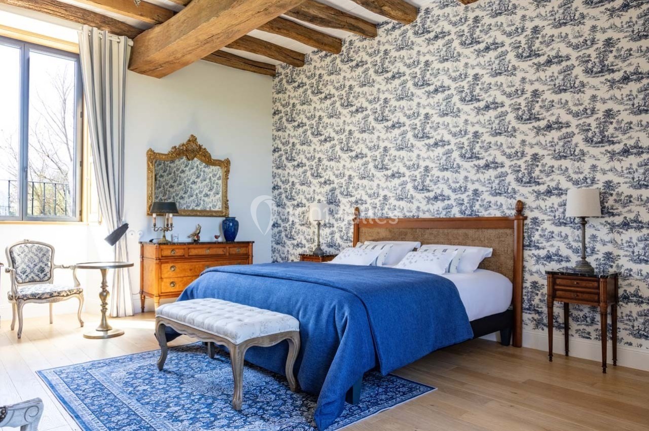 Chambre lumineuse avec lit double, couvre-lit bleu, mobilier en bois, miroir doré et mur décoré de papier peint à motifs.