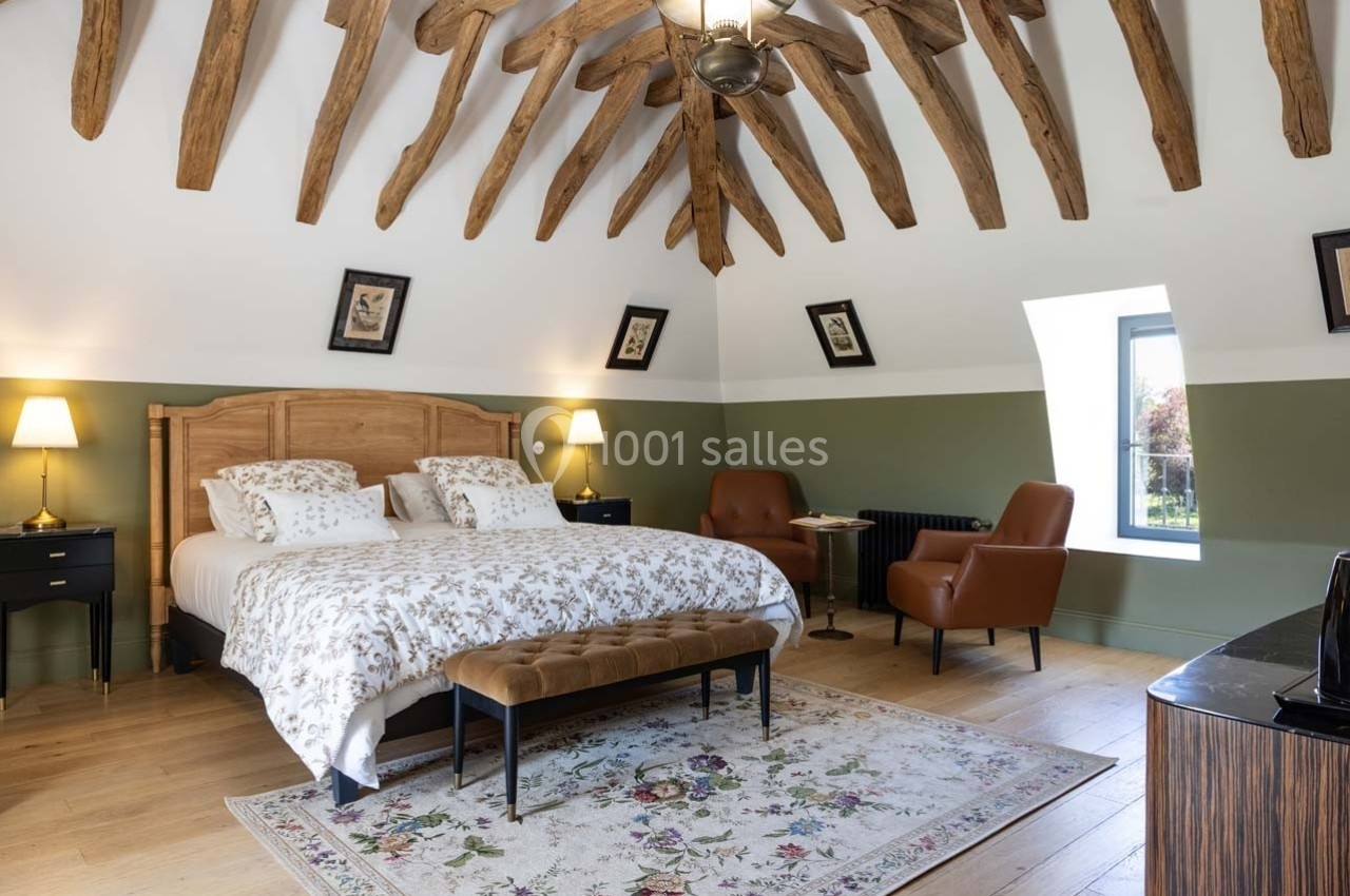 Chambre lumineuse avec poutres apparentes, lit double, fauteuils en cuir, tapis fleuri et vue sur l'extérieur.