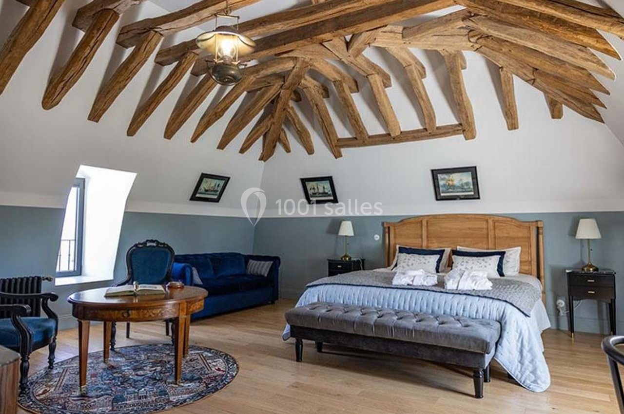 Chambre spacieuse avec poutres apparentes, lit double, canapé, table ronde et décoration classique.